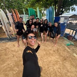 Un groupe WeRoad se prend en selfie sur une plage de sable devant des râteliers de planches de surf colorées.