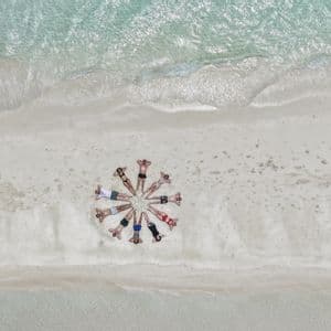 Vue aérienne d'un voyage en groupe WeRoad allongé en cercle sur une plage de sable blanc à côté d'une eau turquoise claire.