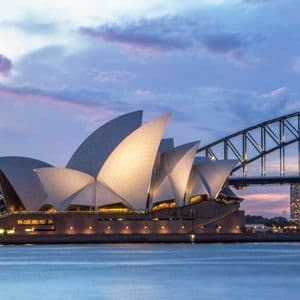 Un edificio moderno con tetti bianchi a forma di vela si erge illuminato accanto a un grande ponte ad arco sull'acqua al tramonto.