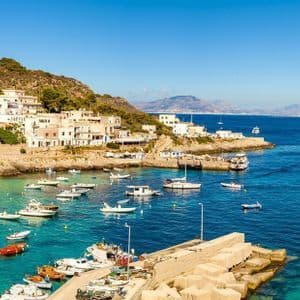 Un village côtier sur une colline verdoyante surplombe une baie remplie de petits bateaux sur une eau turquoise et bleue sous un ciel clair.