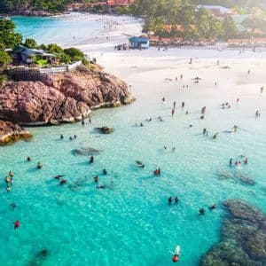 Una vista aerea di persone che nuotano e fanno snorkeling in acque cristalline color turchese vicino a una spiaggia di sabbia bianca sostenuta da rigogliose colline verdi.