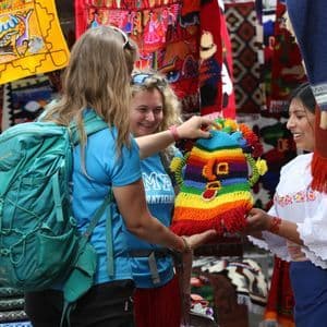 Deux femmes lors d'un voyage en groupe WeRoad regardent un article tricoté coloré tenu par un vendeur local sur un marché en plein air.