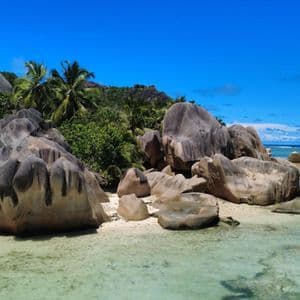 Enormi massi erosi si adagiano su una spiaggia sabbiosa bagnata da acque limpide e turchesi, con palme e una vegetazione lussureggiante su una collina sullo sfondo.