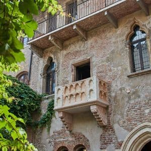Un balcone in pietra ornato si estende da un edificio in mattoni anticato con finestre ad arco, incorniciato da foglie verdi in primo piano.