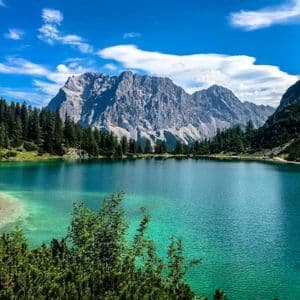 Un lago alpino turchese circondato da una pineta verde, alla base di una grande montagna rocciosa sotto un cielo azzurro.