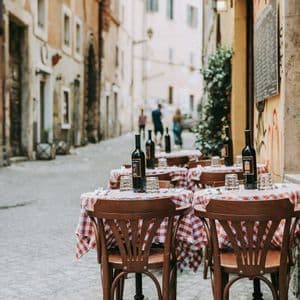 Tavoli con tovaglie a quadretti bianchi e rossi sono apparecchiati con bottiglie di vino in un caffè all'aperto su una strada acciottolata.