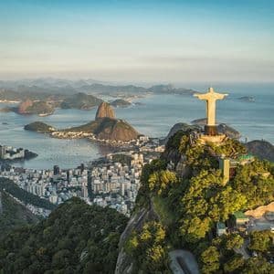 The Christ the Redeemer statue on a mountaintop overlooks a vast coastal city, a bay, and Sugarloaf Mountain at sunrise.
