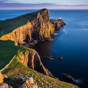 Un faro blanco se asienta en una península rocosa y cubierta de hierba que se adentra en el tranquilo mar azul, iluminado por la cálida luz del atardecer.