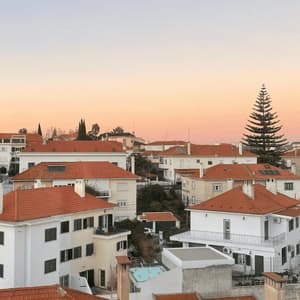 A dense neighborhood of white houses with red-tiled roofs sits on a hillside against a soft, pastel sunset sky.