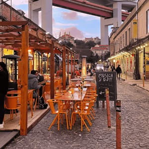 An outdoor restaurant with bright orange chairs is set up on a cobblestone street under a large bridge at dusk.