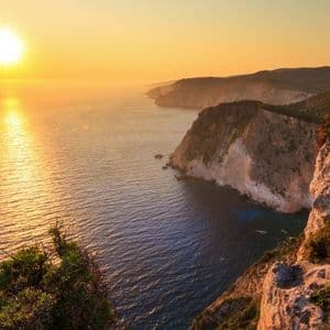 Il sole tramonta sull'oceano, con il suo riflesso che scintilla sull'acqua accanto a una costa rocciosa e ricoperta di arbusti.