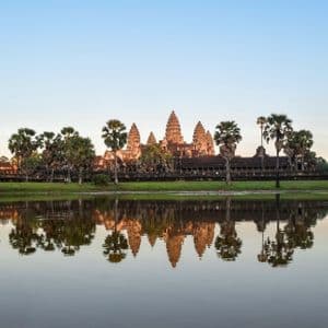 Un grand complexe de temples en pierre et des palmiers environnants se reflètent parfaitement dans un lac calme sous un ciel bleu clair.