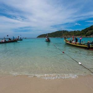 Varias barcas de cola larga con un grupo de viaje de WeRoad flotando en agua turquesa en una playa de arena con una colina boscosa detrás.