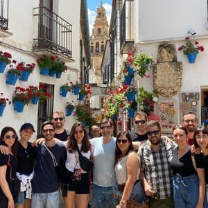 A WeRoad group trip poses for a photo on a narrow street with white walls adorned with blue flower pots and a bell tower in the background.