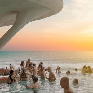 Un gruppo WeRoad che socializza a un moderno swim-up bar in una piscina a sfioro con vista sull'oceano al tramonto.