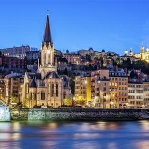 Un paysage urbain au crépuscule avec une église illuminée et des bâtiments le long d'une rivière, avec des lumières se reflétant sur l'eau.