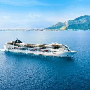 Una grande nave da crociera bianca naviga su un oceano blu calmo con una costa montuosa distante sullo sfondo.