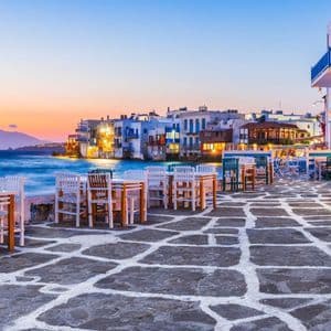 Tavoli e sedie vuoti su un patio acciottolato con vista sul mare e su una città costiera al tramonto.