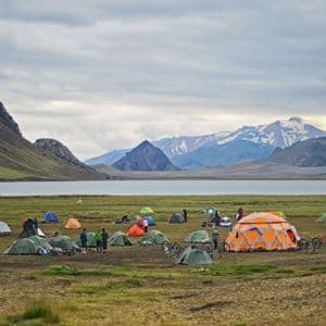 Un voyage de groupe WeRoad avec des tentes colorées et des vélos, campant au bord d'un lac au pied de montagnes escarpées et enneigées.