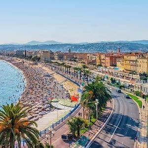 An aerial view of a crowded beach beside a sparkling blue sea, a coastal road, and a city skyline under a clear sky.