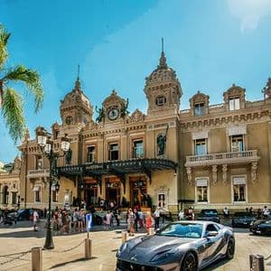 Luxury cars are parked in a plaza in front of an ornate, classical-style building under a clear blue sky.