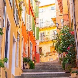 A narrow stone staircase ascends through an alleyway lined with colorful yellow and orange buildings and potted plants.