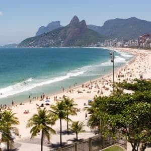 An elevated view of a crowded sandy beach with people swimming, backed by a city and large mountains under a blue sky.