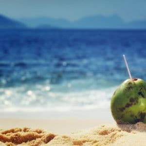 A green coconut with a straw sits on a sandy beach, overlooking the blue ocean and distant mountains.
