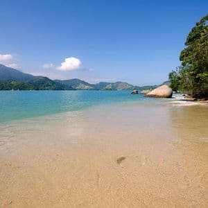 A sandy beach with clear, shallow water leading to a bay with green mountains in the distance under a blue sky.