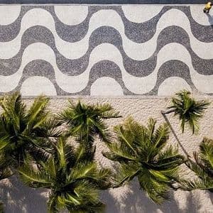 A top-down aerial view of a wavy black-and-white patterned sidewalk next to the tops of palm trees on a sandy beach.