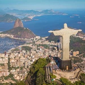 The Christ the Redeemer statue, viewed from behind, overlooks a sprawling cityscape, a bay, and Sugarloaf Mountain.