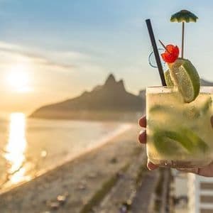 A hand holds a garnished lime cocktail overlooking a beach with mountains in the background at sunset.