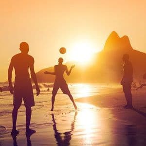 A WeRoad group trip playing with a ball on a beach, silhouetted against a golden sunset with mountains in the background.