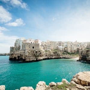 Un borgo con edifici bianchi e in pietra si erge su una scogliera rocciosa a picco su una baia turchese sotto un cielo azzurro parzialmente nuvoloso.