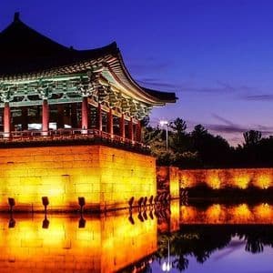 Una tradizionale pagoda asiatica e un muro di pietra illuminati al tramonto, con le loro luci calde che si riflettono nell'acqua immobile.