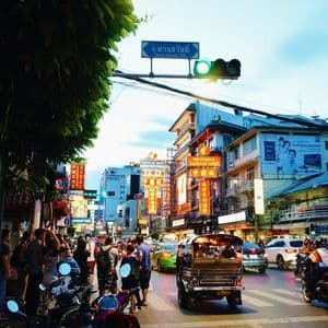 Una concurrida calle de la ciudad al anochecer con un tuk-tuk en un paso de cebra, tráfico, peatones y edificios con letreros de neón brillantes.