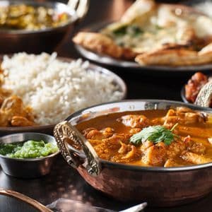 Un gros plan sur une variété de plats indiens, comprenant un poulet au curry dans un bol en cuivre, avec du riz, du chutney et du pain naan en arrière-plan.