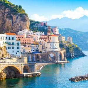 A coastal town with colorful buildings built into a steep cliffside, featuring a stone arched bridge over the blue sea.