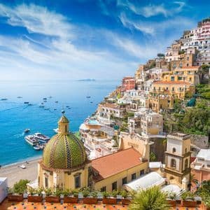 Un pueblo colorido construido en una ladera empinada con una cúpula de azulejos, con vistas a una bahía con barcos bajo un cielo azul.