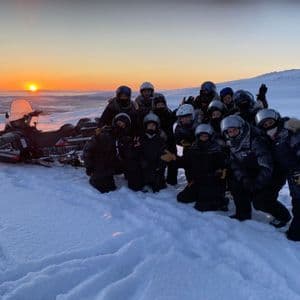 A WeRoad group trip posing for a photo in a snowy landscape next to a snowmobile during sunset.