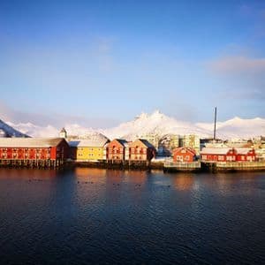 Bunte rot-gelbe Stelzenhäuser säumen einen ruhigen Hafen mit großen, schneebedeckten Bergen im Hintergrund unter blauem Himmel.