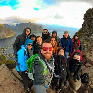 Eine WeRoad-Gruppenreise macht ein Selfie auf einem felsigen Berggipfel, mit Blick auf ein Küstendorf und Berge über eine Bucht.