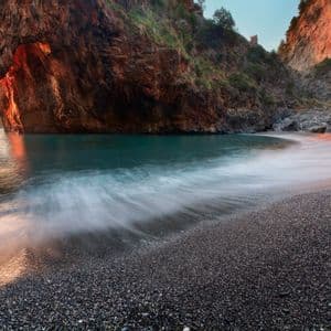 Una foto a lunga esposizione di un'onda che si infrange su una spiaggia di ciottoli scuri in una caletta con un grande arco roccioso illuminato dalla luce del tramonto.
