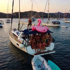 Un viaggio di gruppo WeRoad sorride per una foto su una barca a vela, con un grande fenicottero gonfiabile rosa, circondato da altre barche al tramonto.