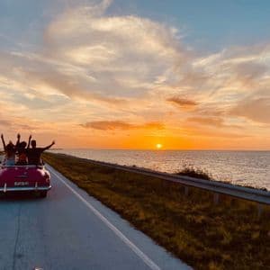Un viaggio di gruppo WeRoad visto da dietro, alla guida di una decappottabile vintage rosa su una strada costiera durante un tramonto dorato.