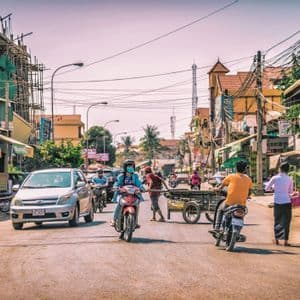 Eine belebte Straßenszene mit Menschen auf Motorrädern, einem Auto und Fußgängern, die an Ladenfronten unter einem Netz von Stromleitungen vorbeiziehen.