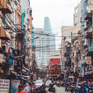 Eine belebte Stadtstraße voller Motorräder und Fußgänger, gesäumt von Gebäuden, einem Gewirr von Oberleitungen und einem modernen Wolkenkratzer im Hintergrund.