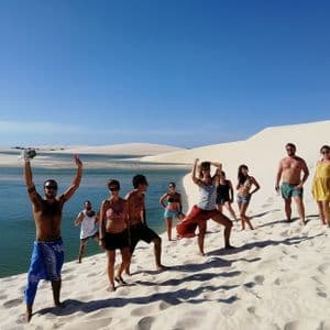 A WeRoad group trip posing on a white sand dune beside a turquoise lagoon under a clear sunny sky.