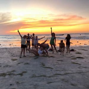 A WeRoad group trip of people posing playfully on a sandy beach with the ocean behind them during a colorful sunset.