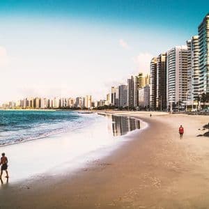 A wide sandy beach with a few people walking along the shoreline, bordered by a modern city skyline with numerous high-rise buildings.